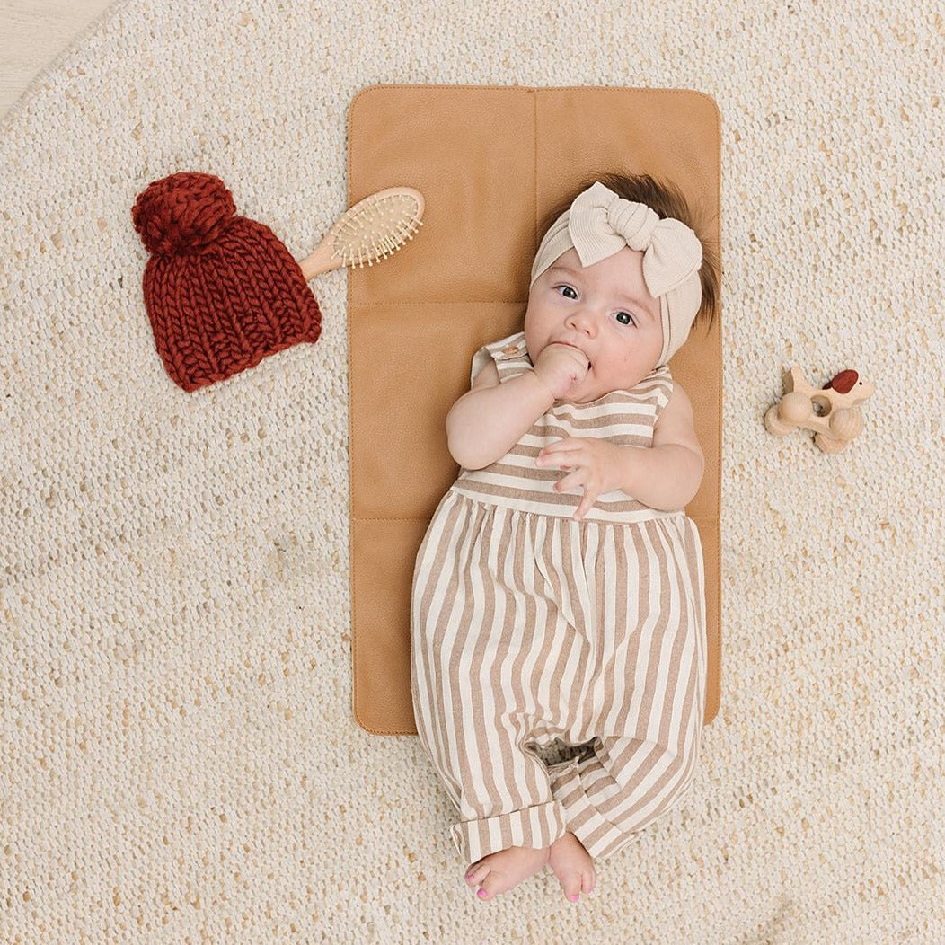 A caregiver holds a baby's hands as the little one, dressed in a blue outfit with a pink bow, rests on a vegan leather Changing Mat placed on a patterned rug.