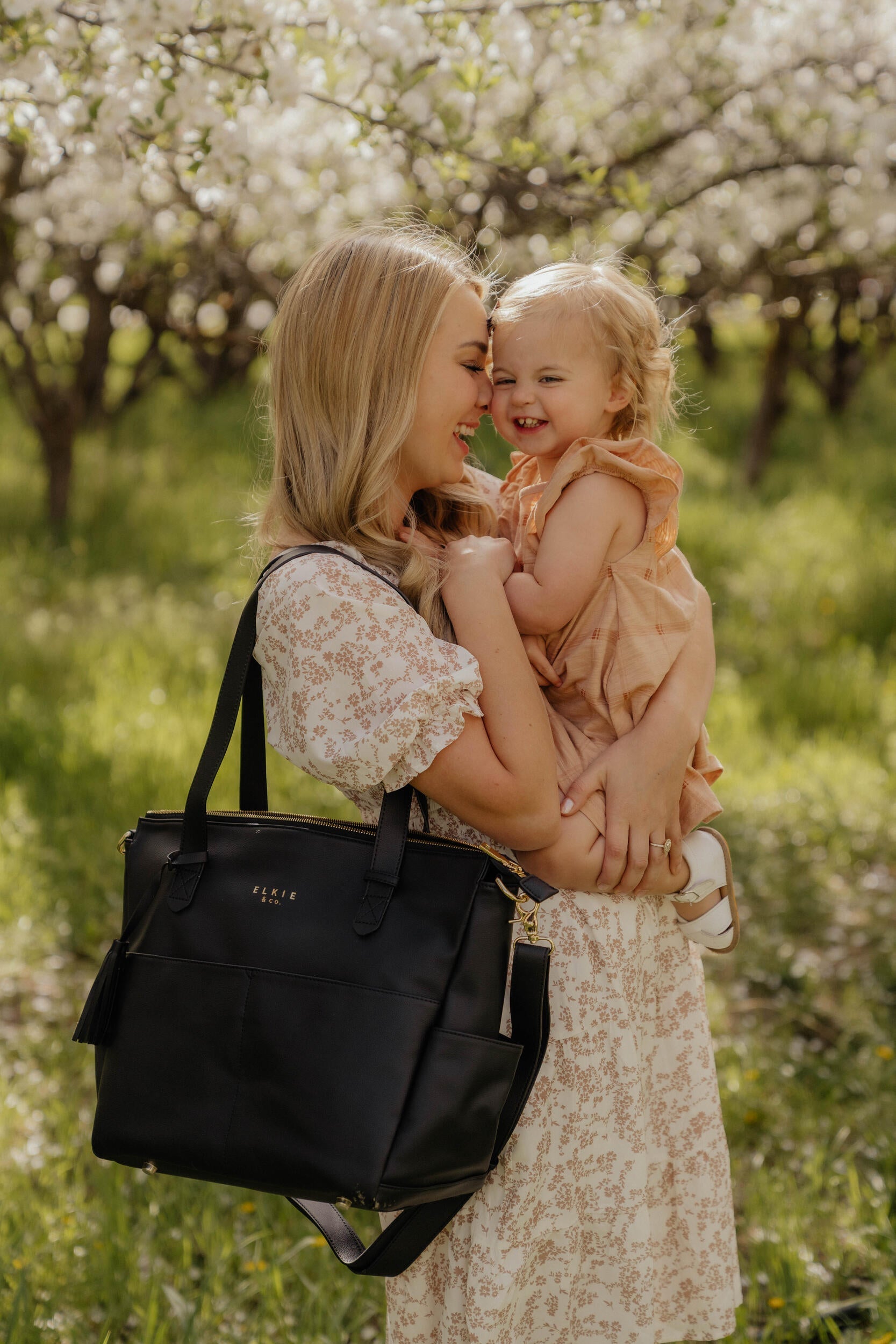 A person stands against a white brick wall, showcasing *The Aberdeen*, a stylish brown vegan leather tote with two front pockets and tassels.