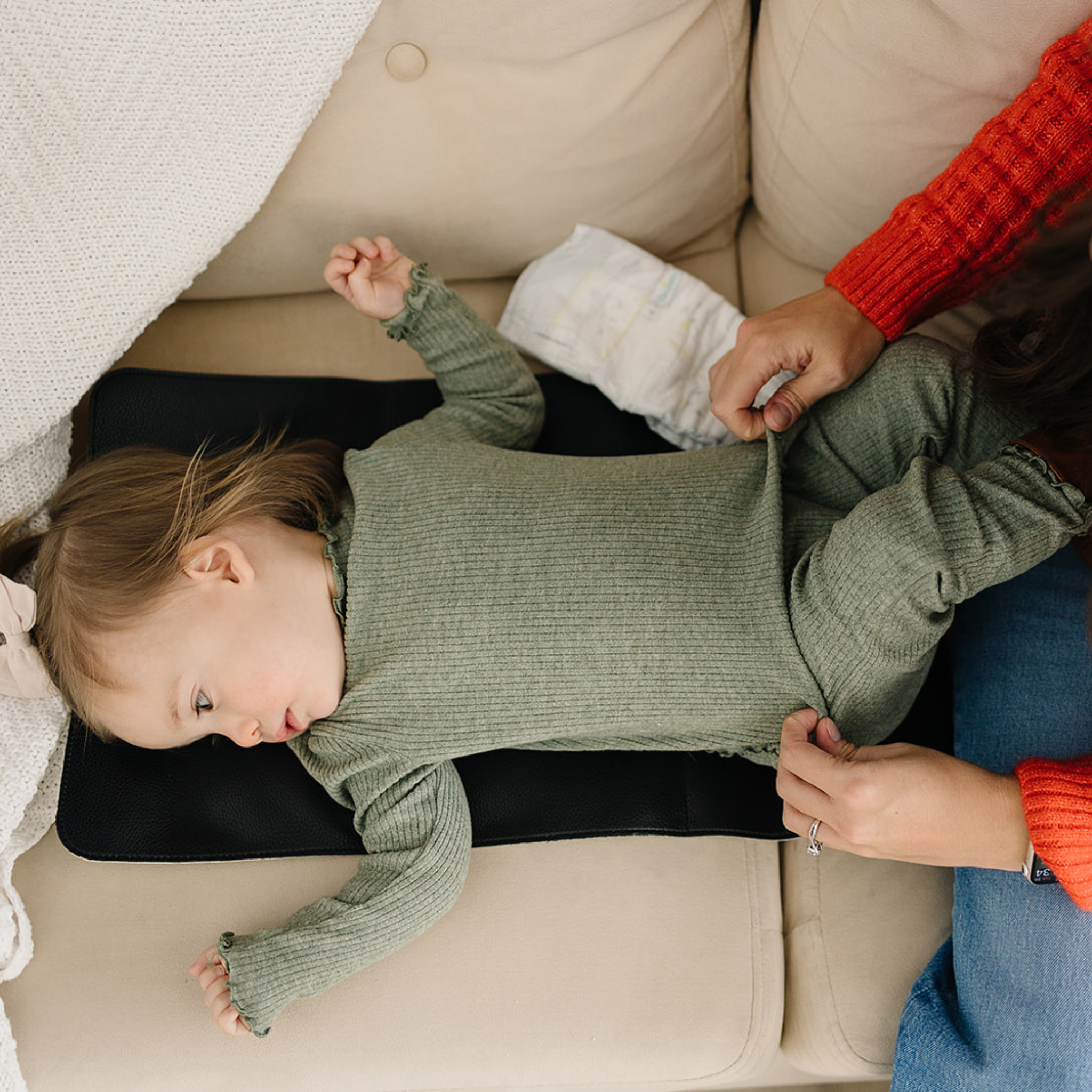 A caregiver holds a baby's hands as the little one, dressed in a blue outfit with a pink bow, rests on a vegan leather Changing Mat placed on a patterned rug.