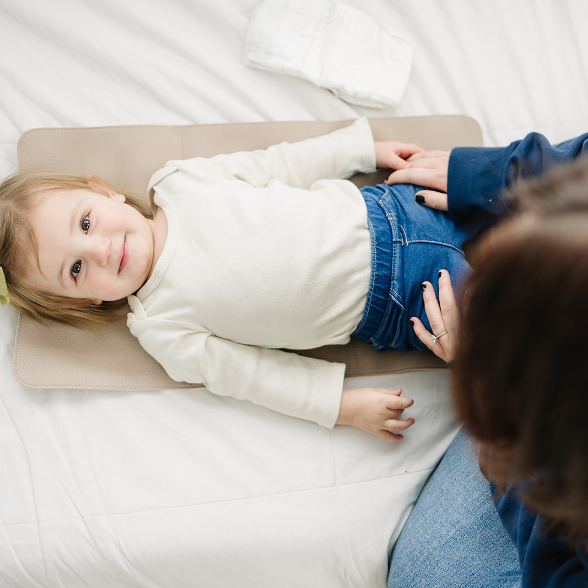 A caregiver holds a baby's hands as the little one, dressed in a blue outfit with a pink bow, rests on a vegan leather Changing Mat placed on a patterned rug.