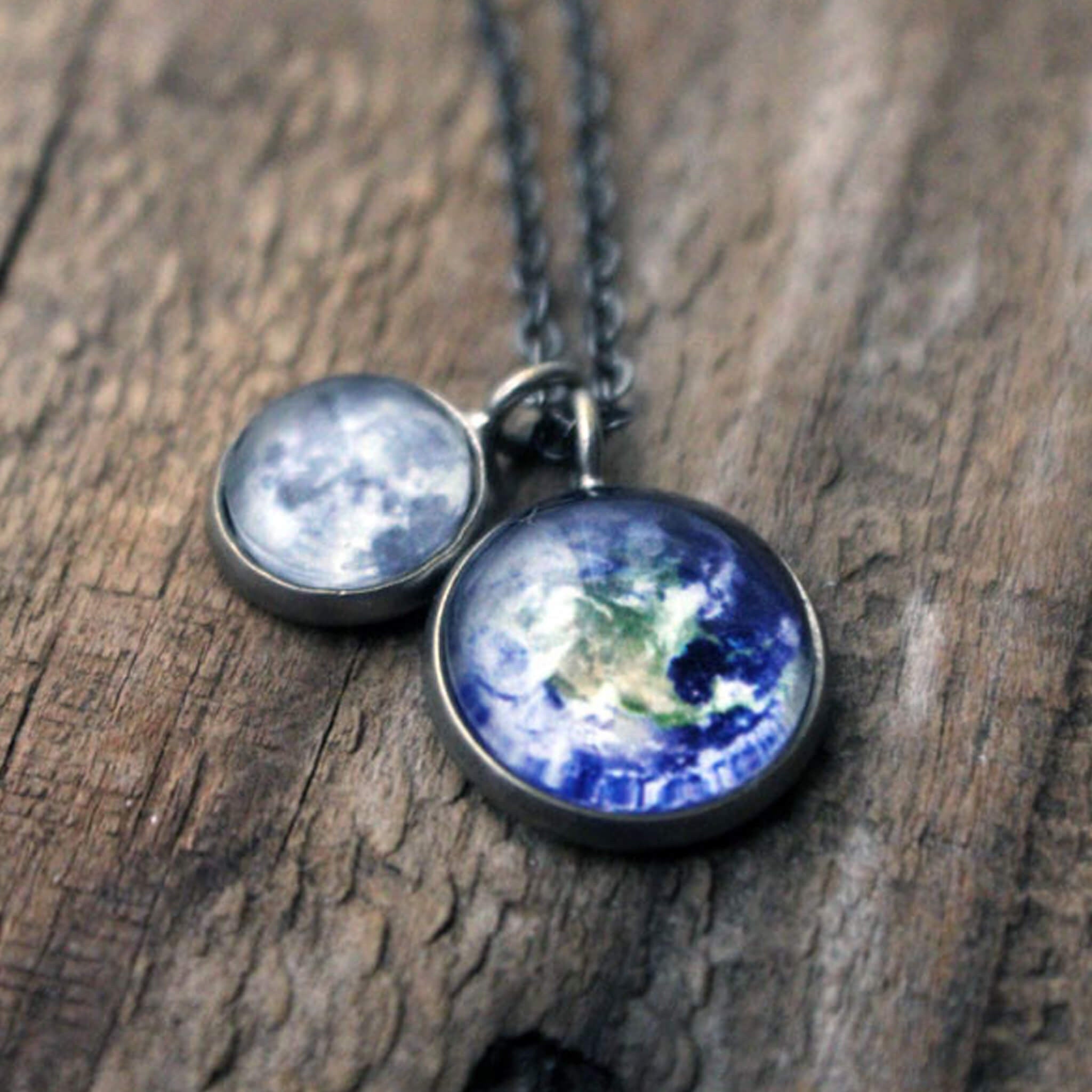 On a wooden surface, two silver Earth and Moon Layered Space Necklaces are adorned with pendants depicting Earth and the Moon. Encircled by small star-shaped embellishments, this celestial jewelry captures the ethereal beauty of our universe.