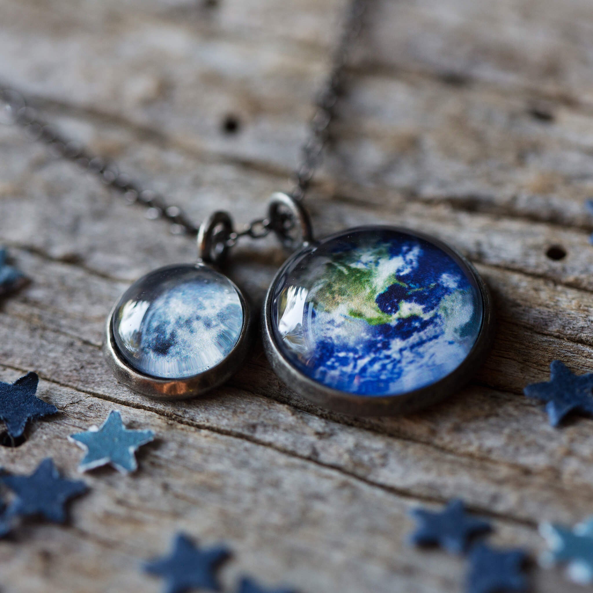 On a wooden surface, two silver Earth and Moon Layered Space Necklaces are adorned with pendants depicting Earth and the Moon. Encircled by small star-shaped embellishments, this celestial jewelry captures the ethereal beauty of our universe.