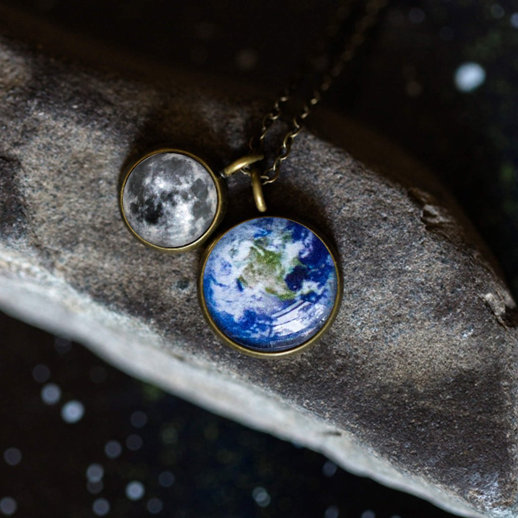 On a wooden surface, two silver Earth and Moon Layered Space Necklaces are adorned with pendants depicting Earth and the Moon. Encircled by small star-shaped embellishments, this celestial jewelry captures the ethereal beauty of our universe.