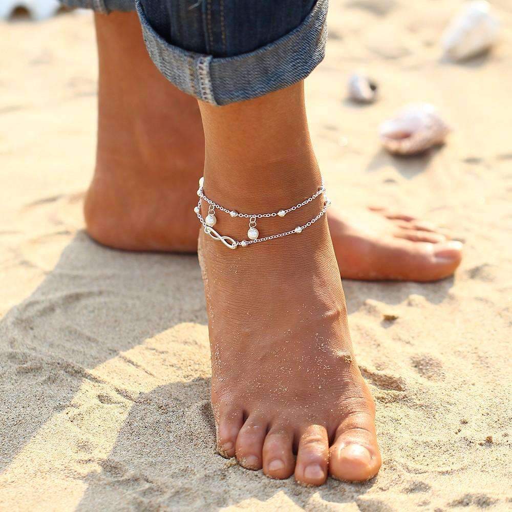 A foot partially covered in sand on the beach is adorned with an Infinity Pendent Pearl Anklet, crafted from zinc alloy with a double layer and featuring an infinity symbol and simulated pearls.