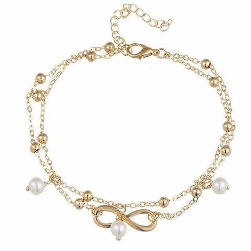 A foot partially covered in sand on the beach is adorned with an Infinity Pendent Pearl Anklet, crafted from zinc alloy with a double layer and featuring an infinity symbol and simulated pearls.