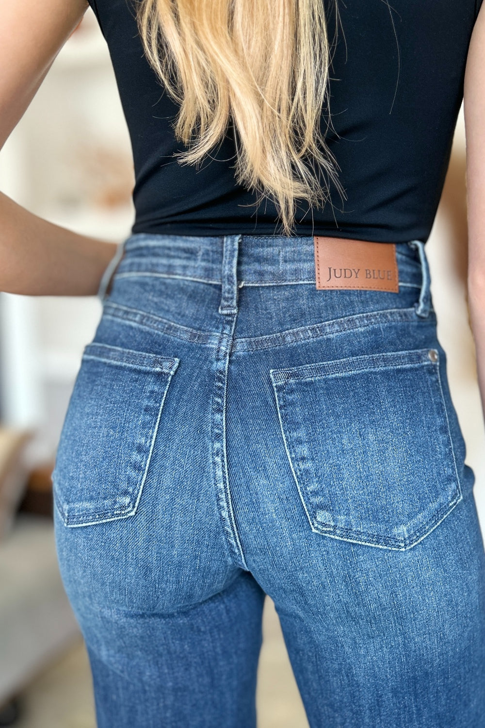 A person wearing a black top and Judy Blue Full Size Tummy Control Straight Jeans stands indoors with one hand on their hip. Their shoes are black with white soles. A plant and shelves are in the background.