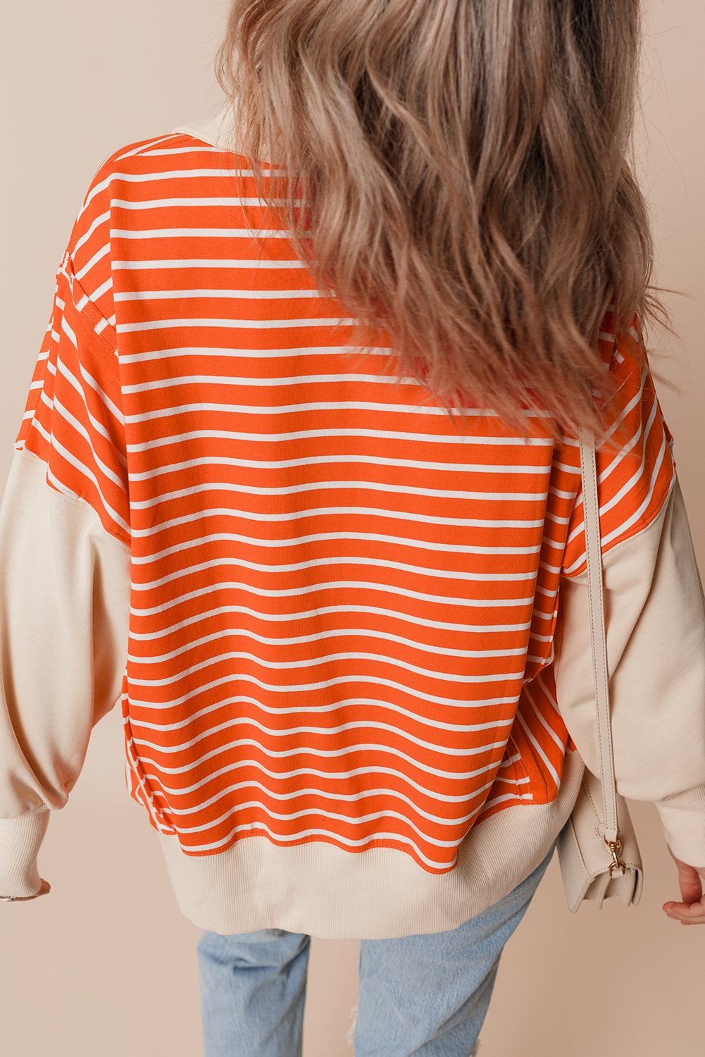 A person with wavy hair is seen from the back, wearing an Orange Stripe Color Block Loose Fit Collared Drop Shoulder Sweatshirt, light blue jeans, and carrying a small beige bag.