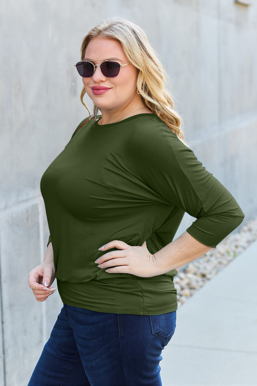 A woman in a slightly stretchy, brown Basic Bae Full Size Round Neck Batwing Sleeve top and blue jeans stands against a concrete wall, holding a brown handbag.