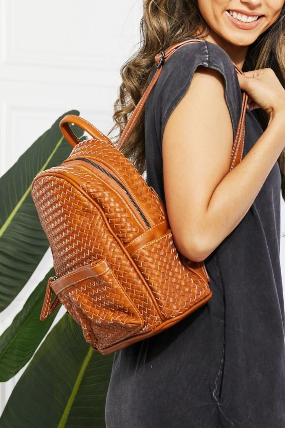 A person wearing the SHOMICO Certainly Chic Faux Leather Woven Backpack over one shoulder, with long hair and a dark shirt, standing sideways.