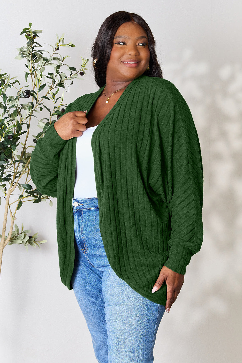 A person with wavy hair, wearing the Basic Bae Full Size Ribbed Cocoon Cardigan over a white top and beige pants, stands in front of a light-colored background with a leafy plant, showcasing an effortlessly basic style.