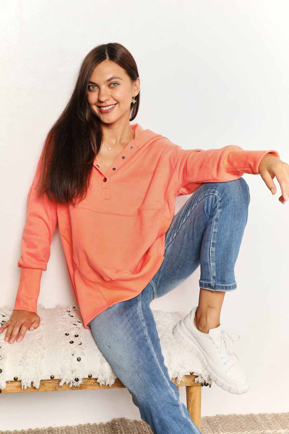 A woman with long hair wears a Double Take Quarter-Snap Dropped Shoulder Hoodie in solid orange. She is standing, with one hand touching her hair and the other resting in the pocket of her jeans, exuding a casual vibe.