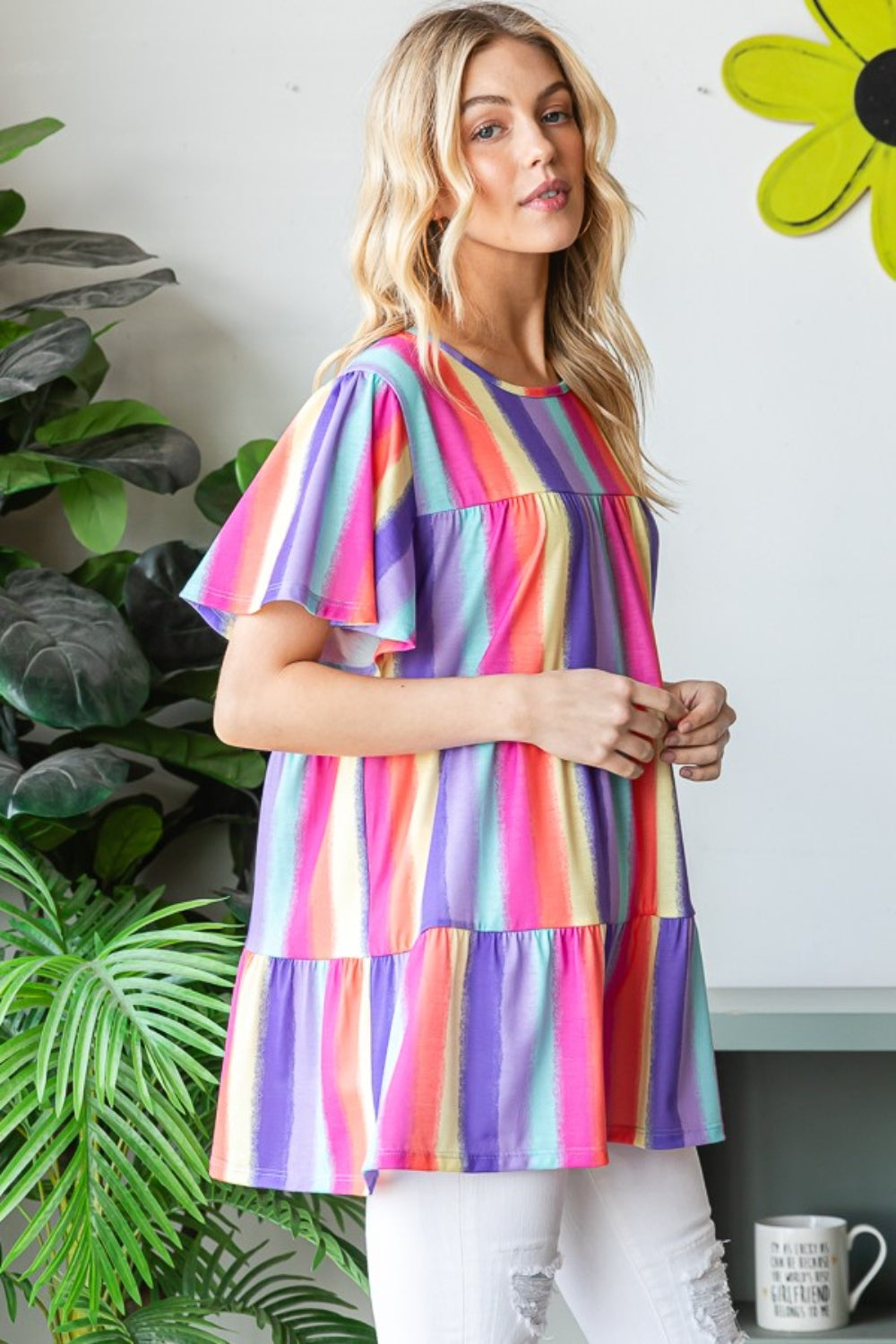 A person wearing the Heimish Full Size Short Sleeve Striped Tiered Top, featuring a colorful, classic striped pattern and flared sleeves, stands by a window. They are smiling, surrounded by plants in the background.