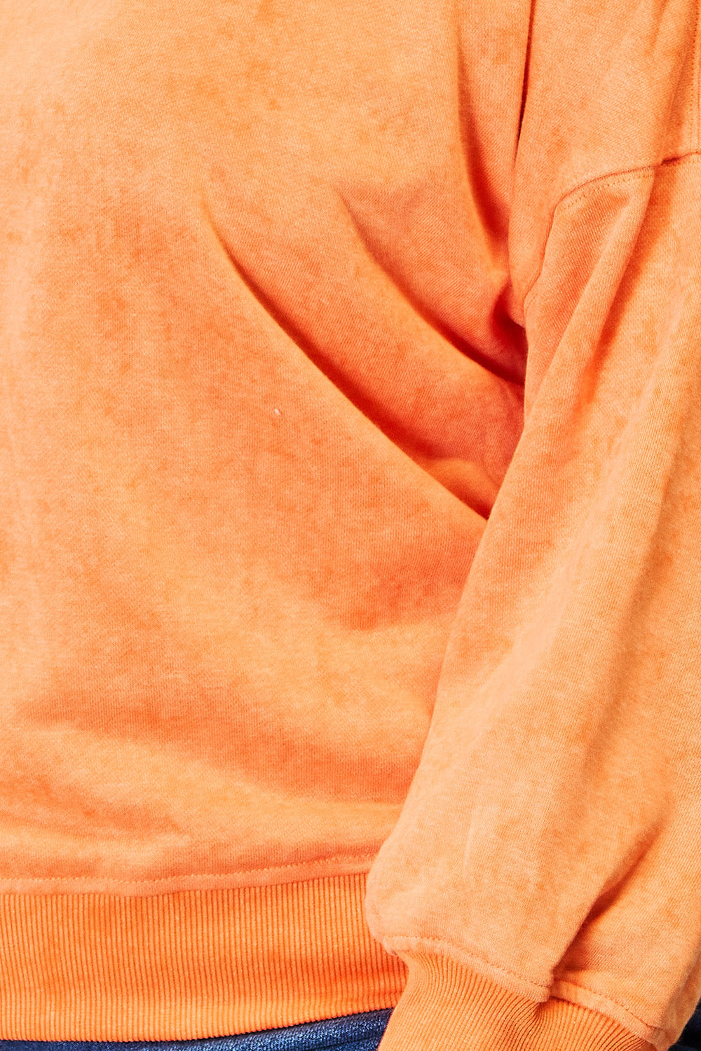 A woman with long, wavy hair smiles in a casual wardrobe, sporting the Basic Bae Round Neck Dropped Shoulder Sweatshirt in orange and blue jeans. She stands beside a white dresser adorned with decorative vases.