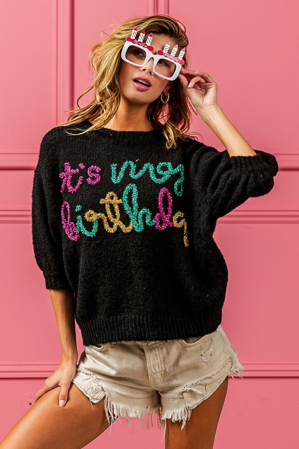 A woman poses against a pink wall, wearing a BiBi Metallic Letter Puff Sleeve Hairy Sweater that reads "It's My Birthday," beige shorts, and novelty glasses with birthday candles.