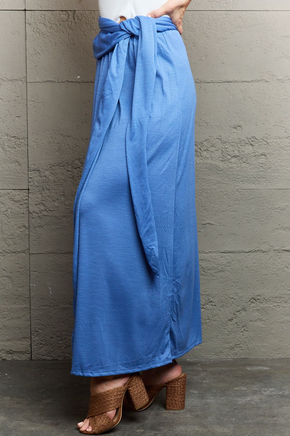 A person stands against a textured gray wall, smiling in the Ninexis Know Your Worth Criss Cross Halter Neck Maxi Dress in blue, featuring a stylish cut-out design. One hand rests on the wall while the other is confidently placed on their hip.