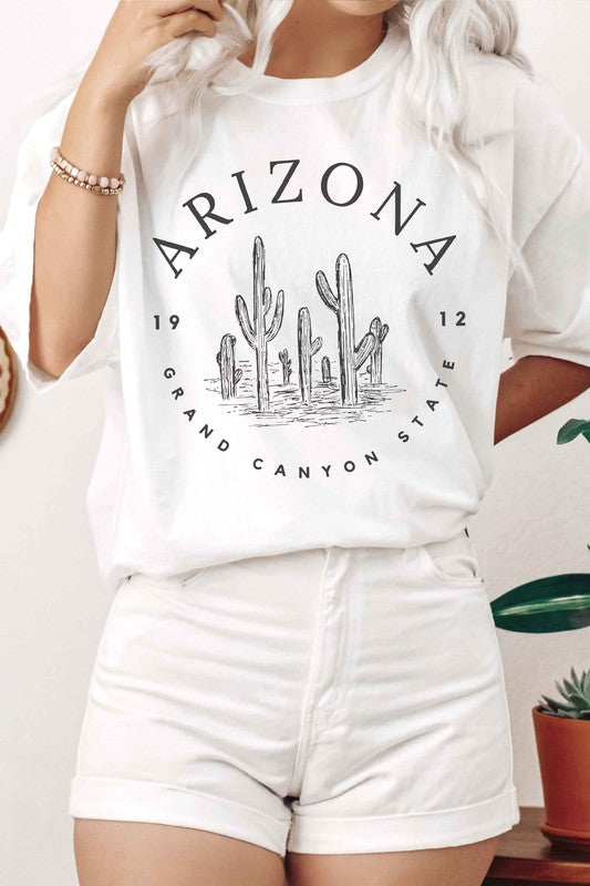 Person wearing an ARIZONA GRAND CANYON STATE Graphic Tee, made of premium 100% cotton with a design featuring the words "ARIZONA GRAND CANYON STATE 1912" and a drawing of cacti, paired with white shorts.