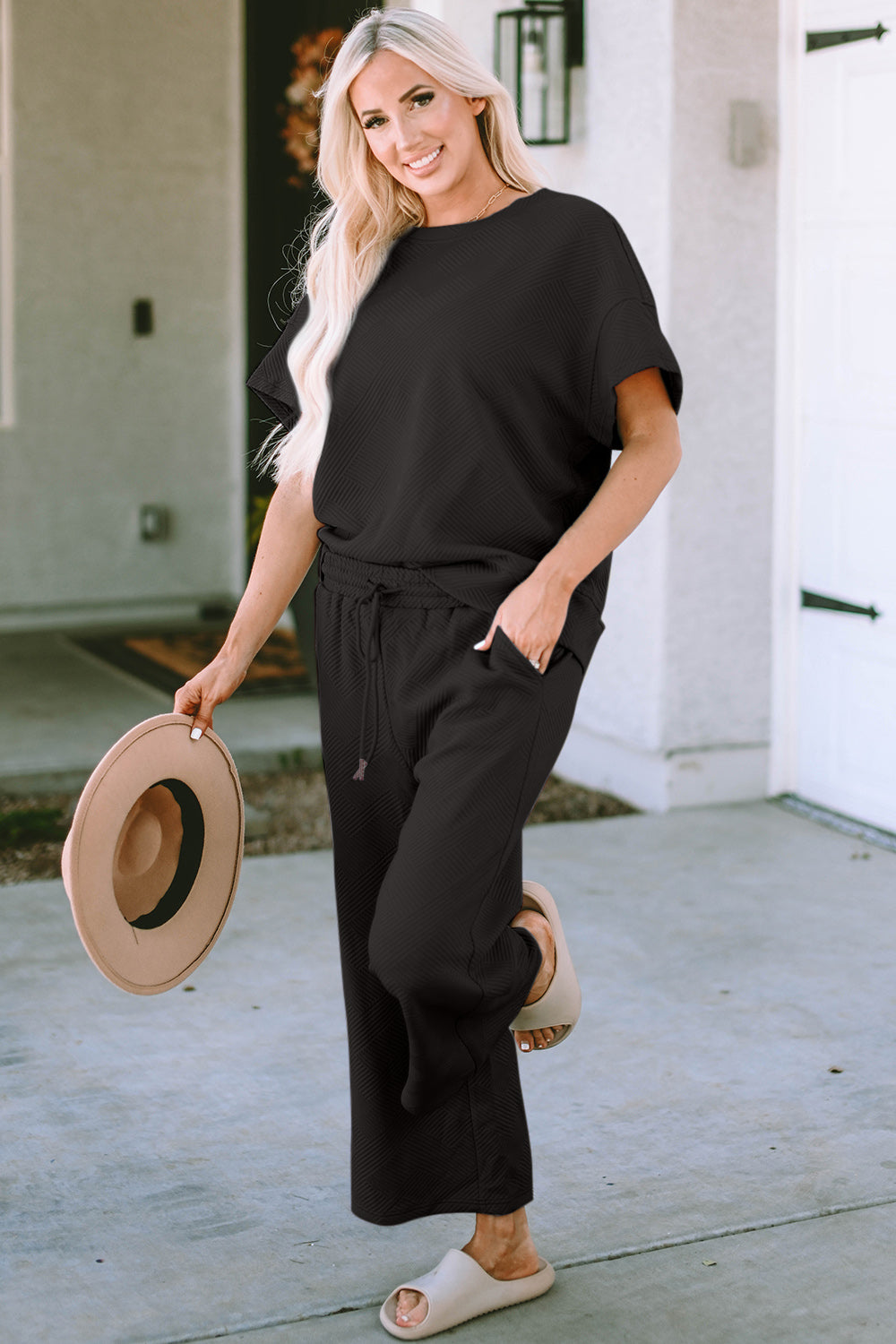 A woman with long hair smiles while standing outside a building, dressed in the Double Take Full Size Texture Short Sleeve Top and Pants Set, featuring a matching beige two-piece ensemble. She wears slides and holds a hat in her right hand. Her outfit, slightly stretchy for added comfort, is perfect for machine wash cold.