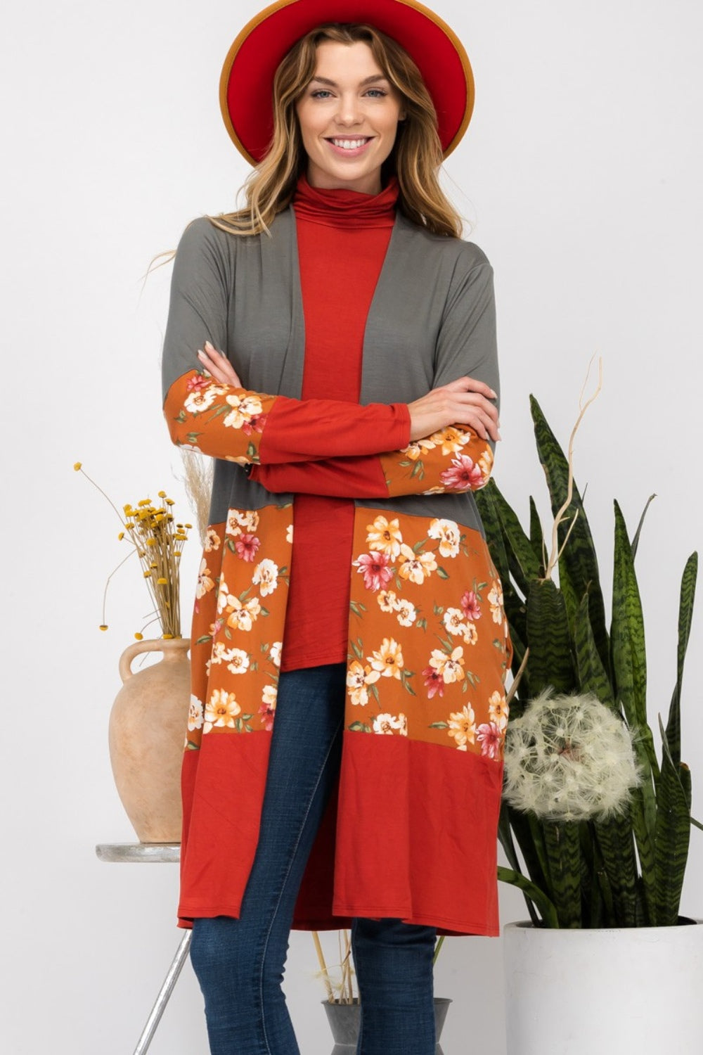 Wearing the Celeste Full Size Floral Color Block Open Front Cardigan, a person in a red hat stands indoors near potted plants, smiling.