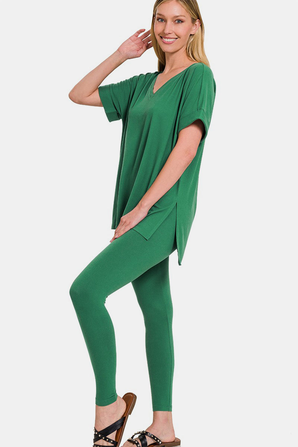 A person is modeling the Zenana Full Size V-Neck Rolled Short Sleeve T-Shirt and Leggings Lounge Set, radiating comfort and style, as they smile and stand with one hand touching their hair.