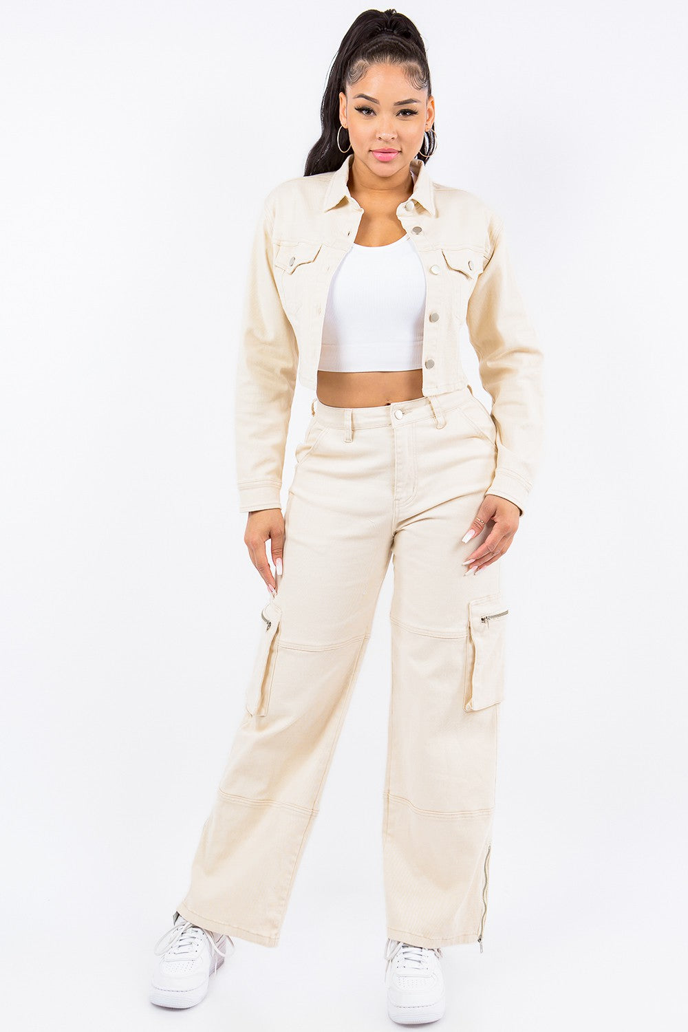 A person wearing an American Bazi Laced Back Cropped Jacket in beige with matching pants and a white crop top stands against a plain white background. They have long dark hair tied in a high ponytail and are looking towards the camera.