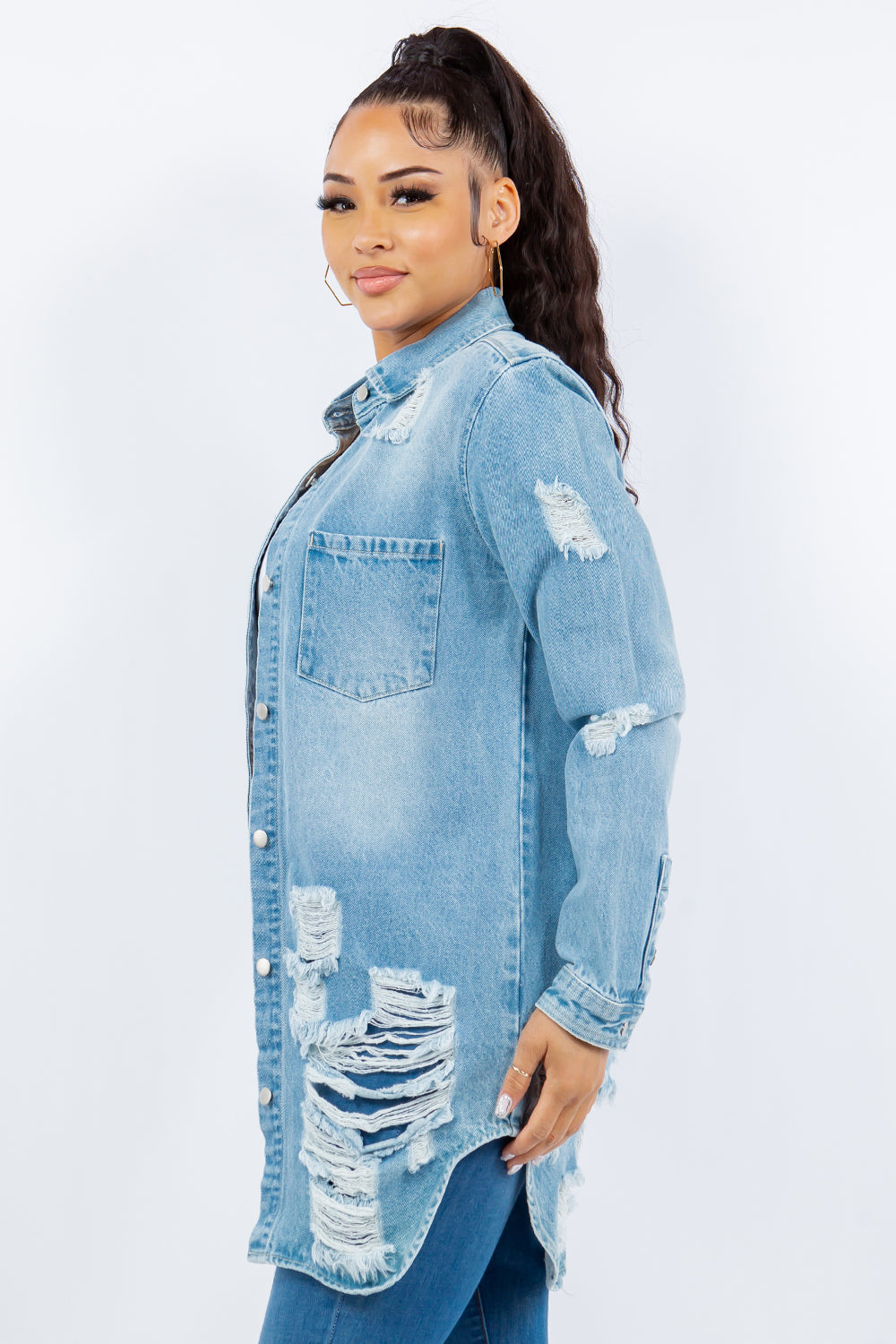 A woman with long hair is wearing an American Bazi Distressed Button Up Long Sleeve Denim Jacket over a white crop top and blue jeans. She is facing forward and standing against a plain white background.