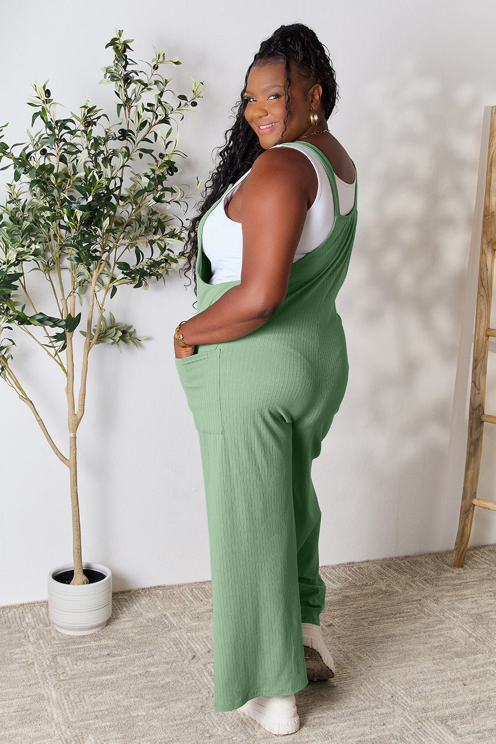 A smiling person wearing the "Double Take Full Size Wide Strap Overall with Pockets," known for its basic style and size-inclusive fit, stands indoors next to a potted plant.