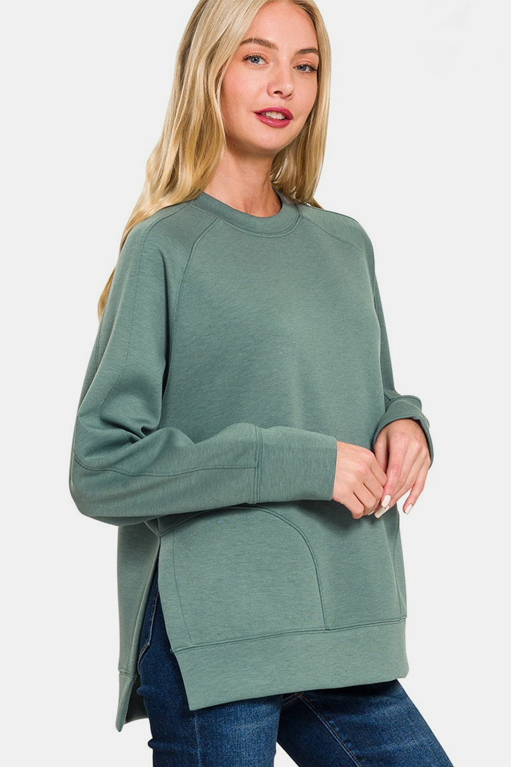 A woman in a Zenana Scuba Round Neck Side Slit Sweatshirt and blue jeans stands against a white background.