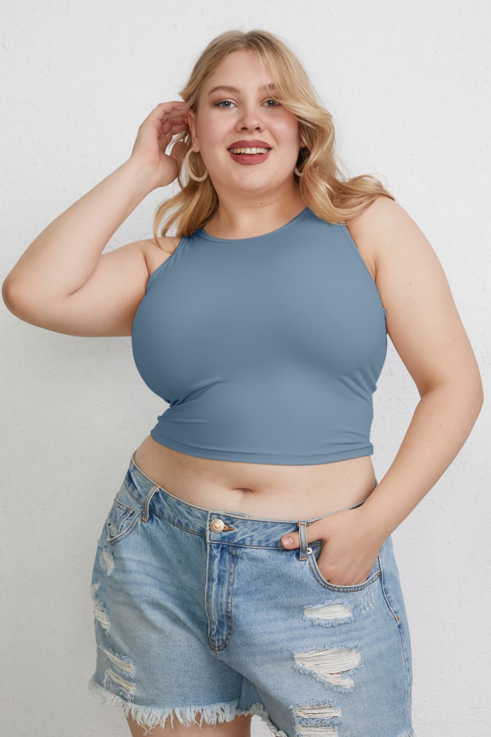 A woman with light hair wears the Basic Bae Full Size Round Neck Slim Tank in black and blue jeans, standing against a light-colored curtain.