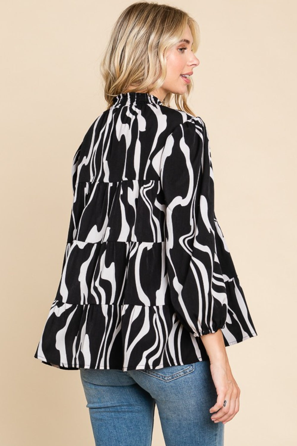 A person wearing a Culture Code Printed Smock Neck Tiered Blouse, featuring a black and white patterned design and long sleeves, paired with blue jeans, stands against a beige background.