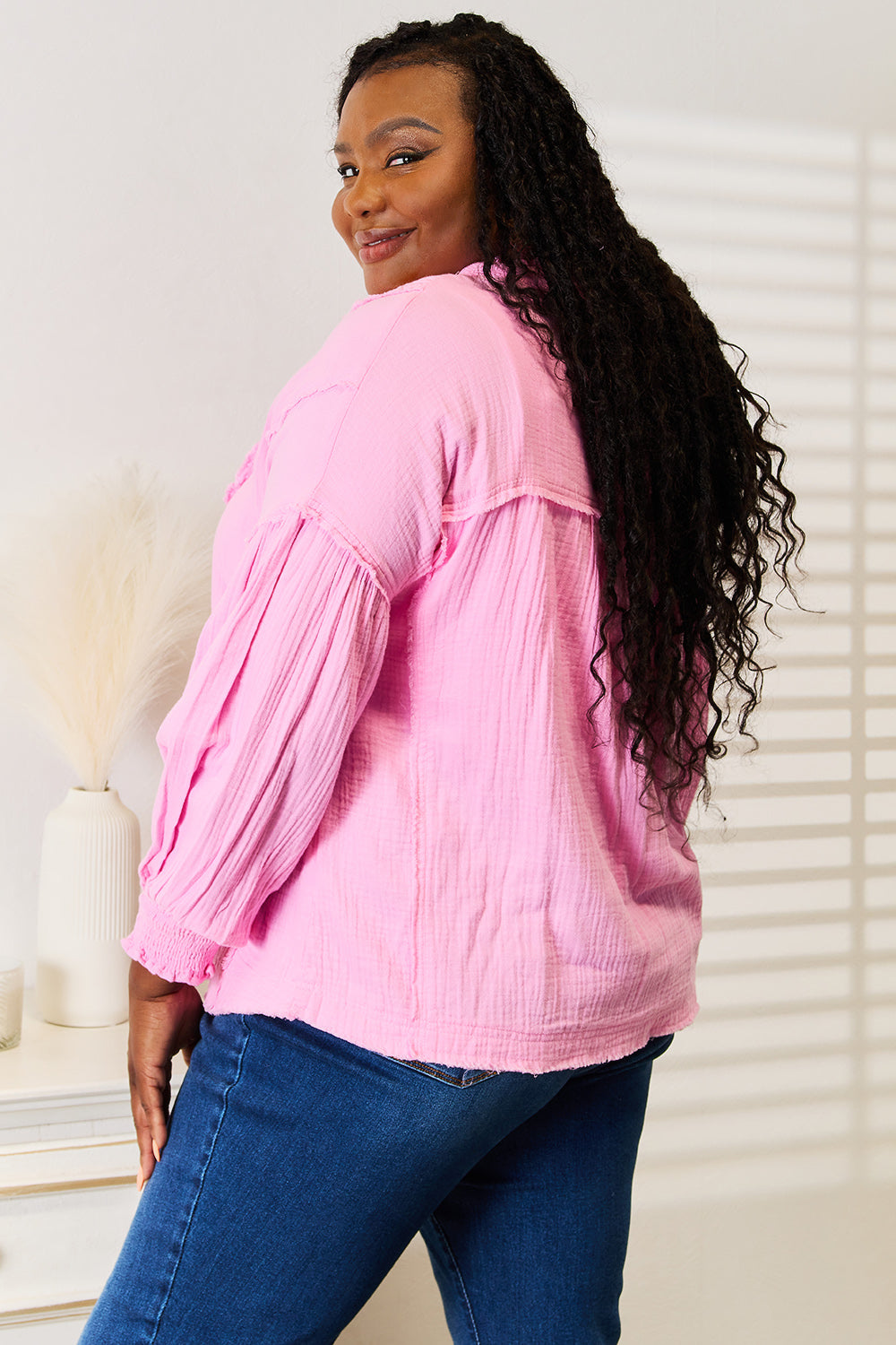 A person wearing a Double Take Exposed Seam Buttoned Notched Neck Blouse in pink and blue jeans touches their ear while standing indoors.