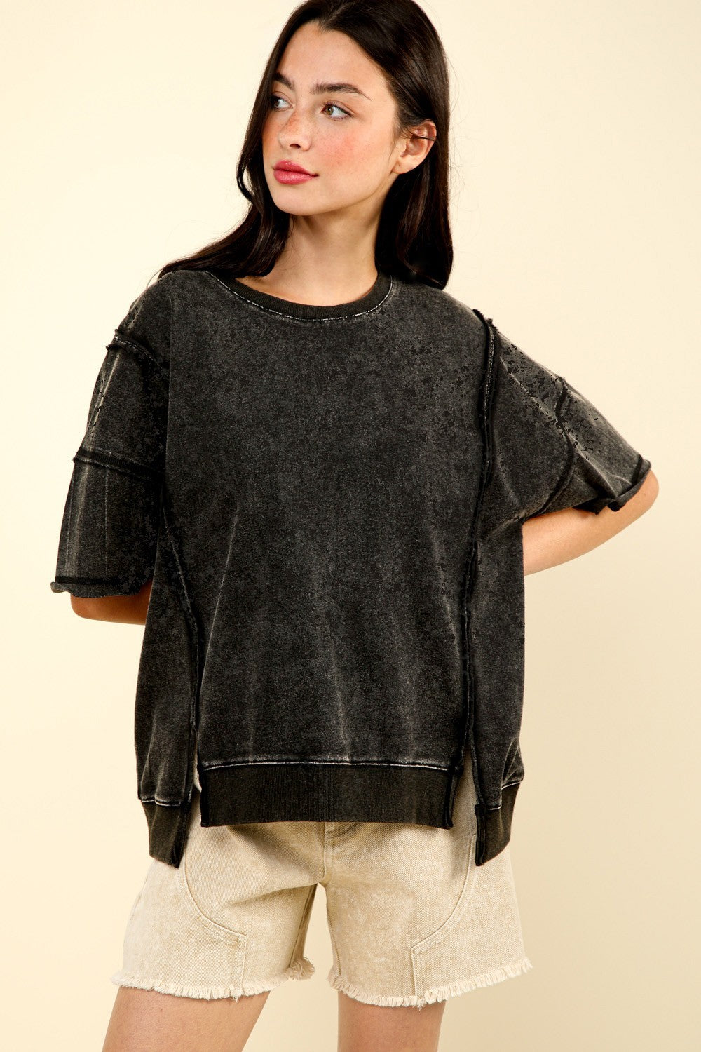 A woman with long dark hair stands against a plain background, wearing an oversized VERY J Round Neck Exposed Seam Slit T-Shirt in dark grey and beige shorts.