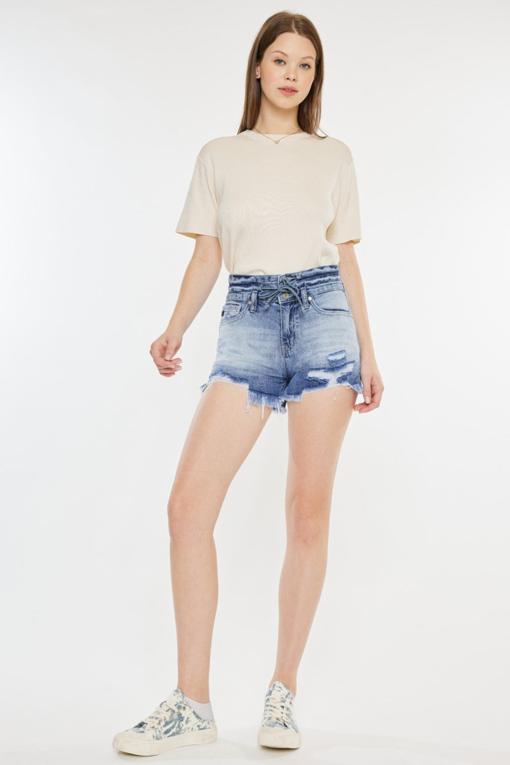 A person wearing a beige t-shirt and Kancan High Rise Frayed Hem Denim Shorts, paired with patterned sneakers, stands against a white background—an ideal addition to any summer wardrobe.