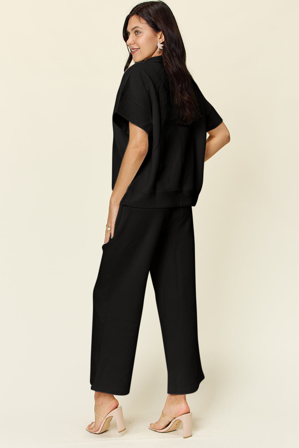 A woman is wearing the Double Take Full Size Texture Half Zip Short Sleeve Top and Pants Set, featuring a white zip-up short-sleeve top and matching wide-leg, drawstring pants. She stands with one hand in her pocket and smiles against a light background.