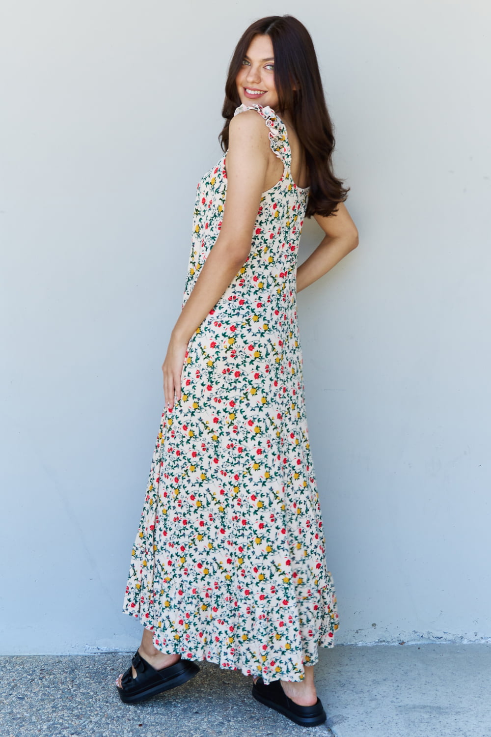 A woman is standing against a gray wall, wearing the Doublju In The Garden Ruffle Floral Maxi Dress in Natural Rose and black sandals, with her left hand raised to her head.