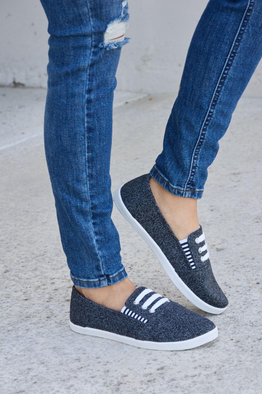 A person wearing ripped blue jeans and the fashionable Forever Link Round Toe Slip-On Flat Sneakers with white elastic bands.