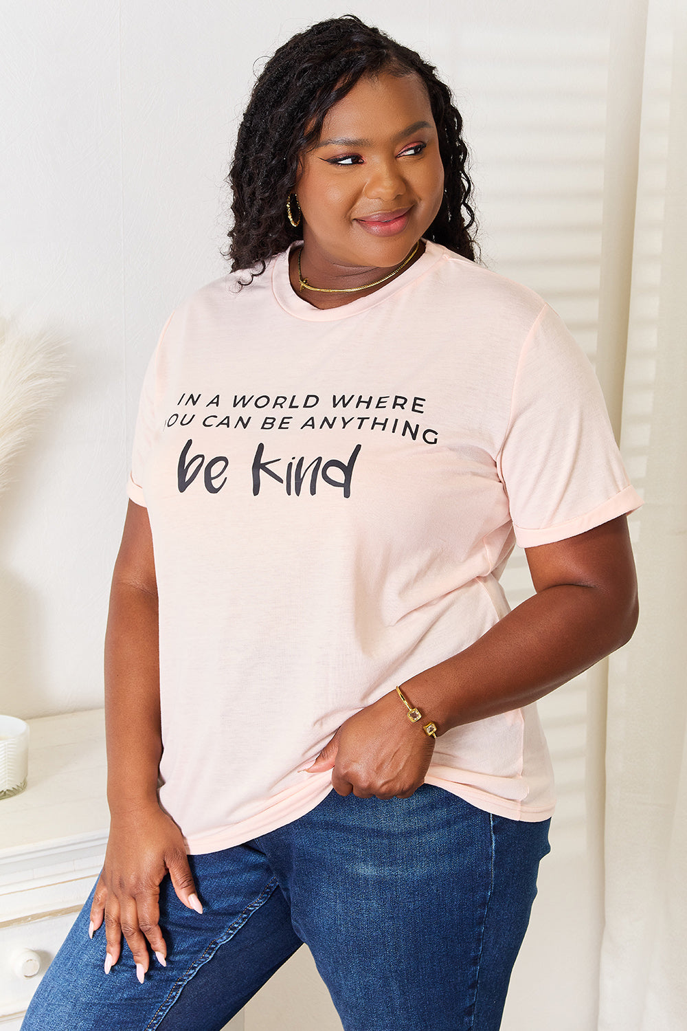 A person wears a Simply Love Slogan Graphic Cuffed T-Shirt in light pink, adorned with the inspiring message, "In a world where you can be anything, be kind," and featuring stylish cuffed sleeves.