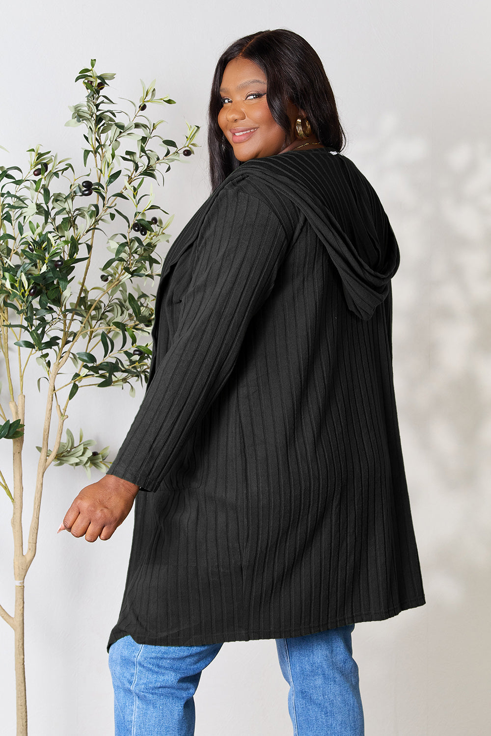 A woman wearing the Basic Bae Full Size Hooded Sweater Cardigan in black, paired with a white top and khaki pants, stands in front of a light background accented by a leafy plant on her left. With one hand tucked into her pocket, she gazes to her right.