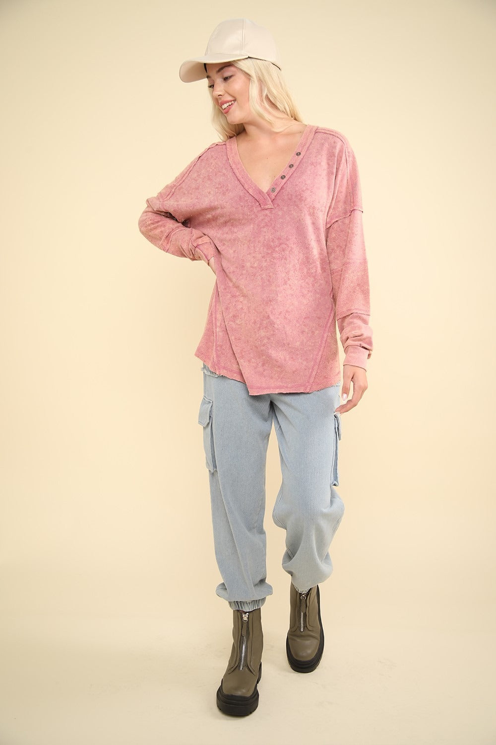 A person wearing a VERY J Washed V-Neck Exposed Seam Knit Top in pink, paired with light blue pants and a beige cap, poses against a beige background.