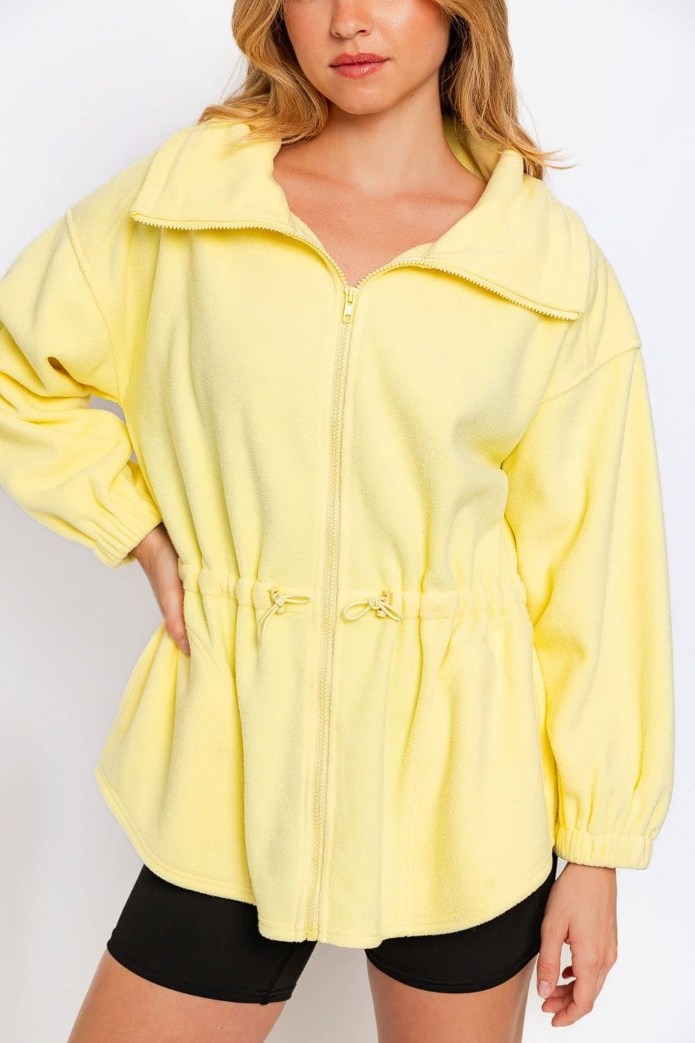 A woman wearing a black outfit and a yellow Le Lis Zip Up Waist Drawstring Soft Fleece Jacket stands against a plain white background.