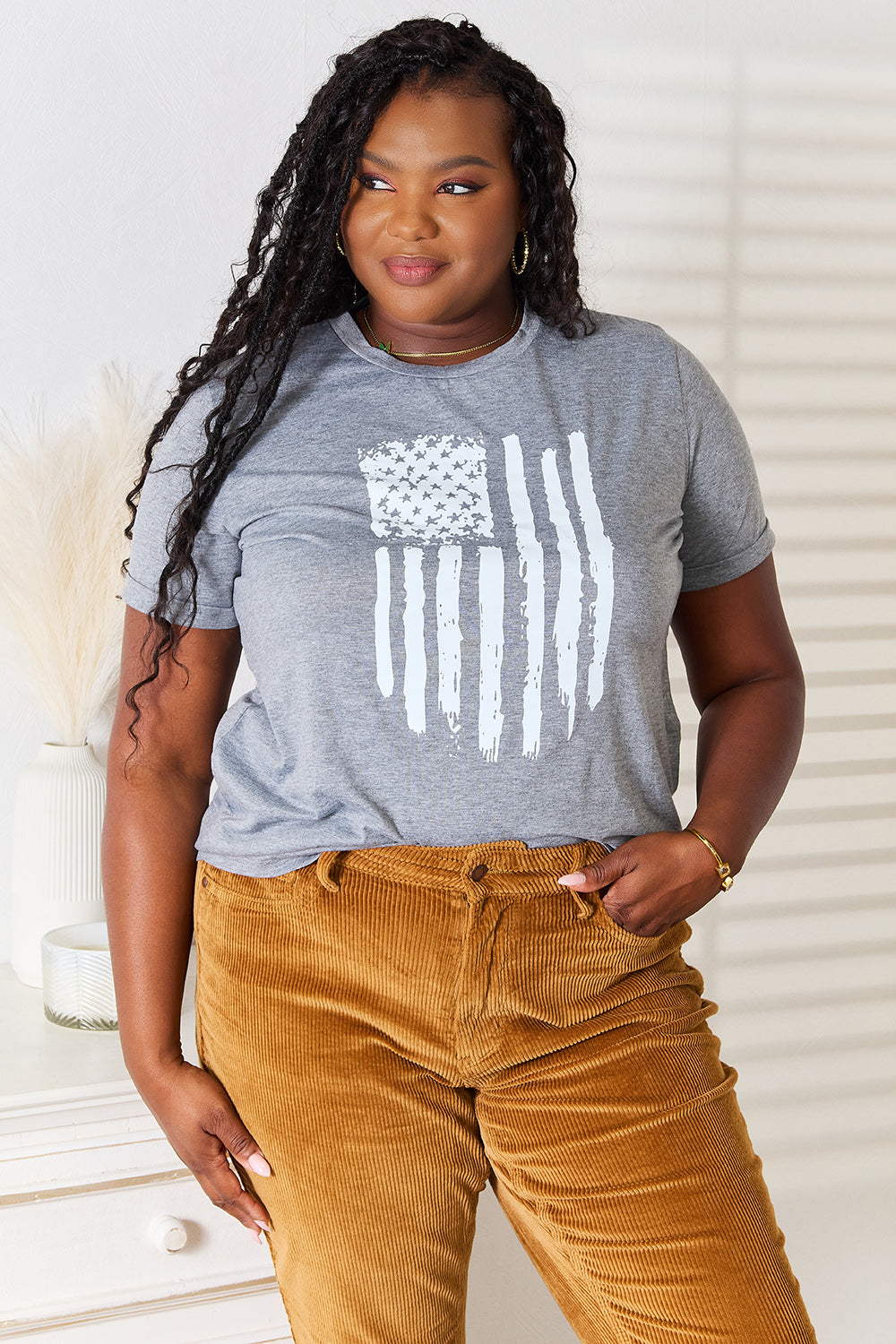 A woman with long dark hair is wearing a Simply Love US Flag Graphic Cuffed Sleeve T-Shirt and brown pants as she stands indoors against a light-colored background.