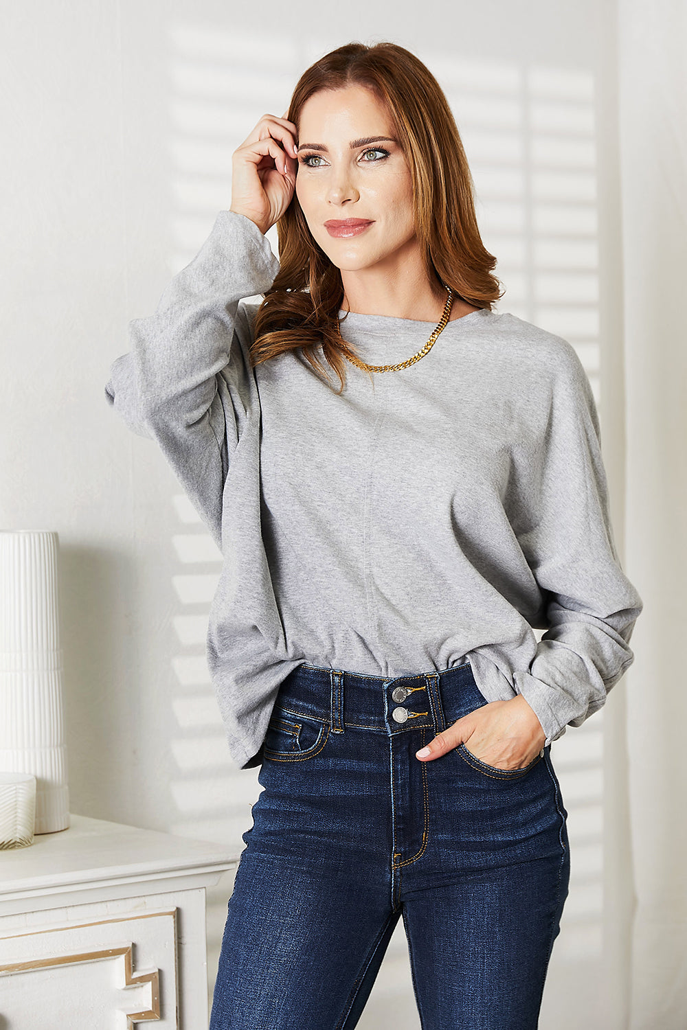 A person with long hair wearing a Double Take Seam Detail Round Neck Long Sleeve Top in solid black and blue jeans stands with one hand in their pocket, looking downward and smiling.