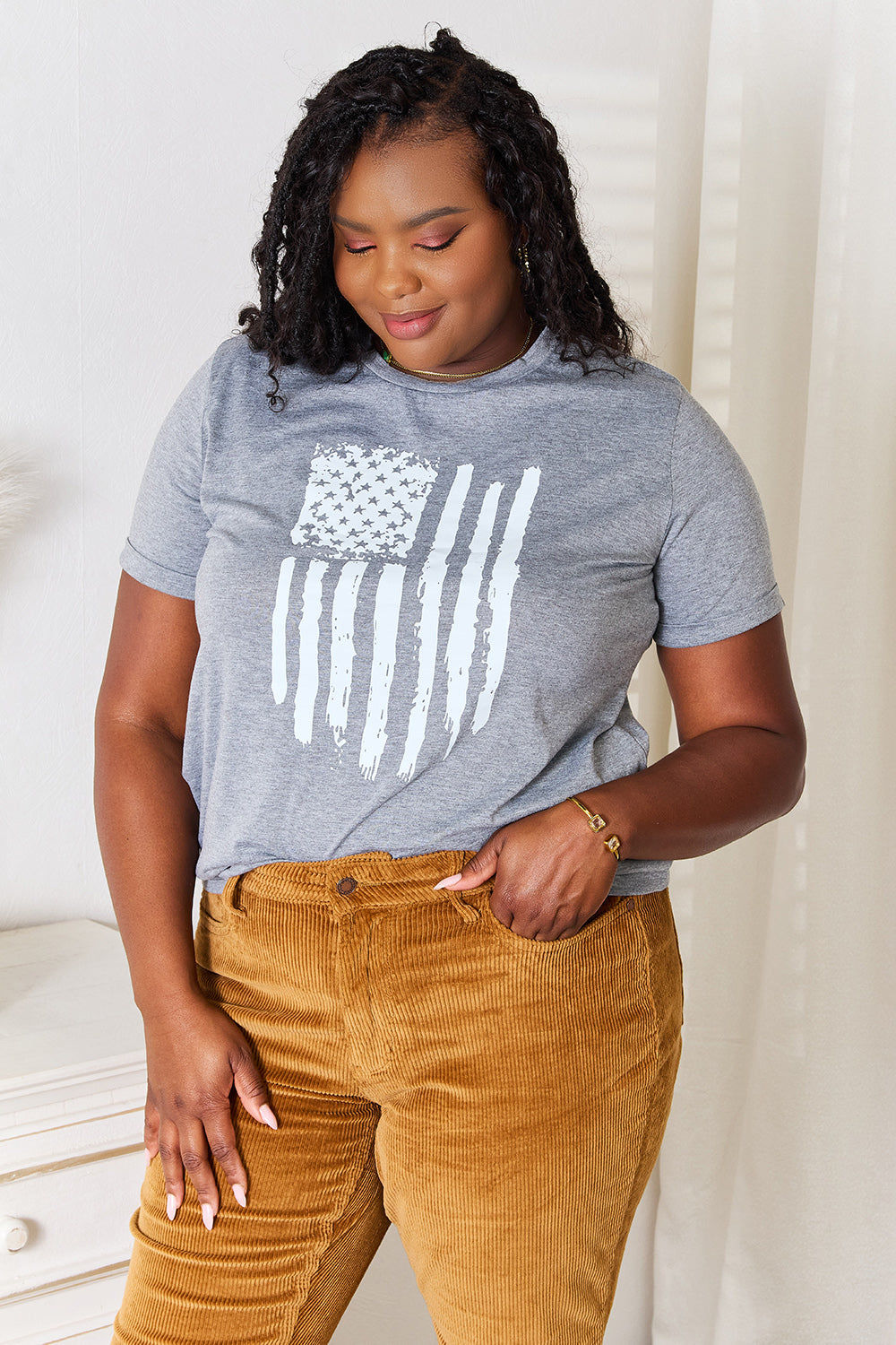 A woman with long dark hair is wearing a Simply Love US Flag Graphic Cuffed Sleeve T-Shirt and brown pants as she stands indoors against a light-colored background.