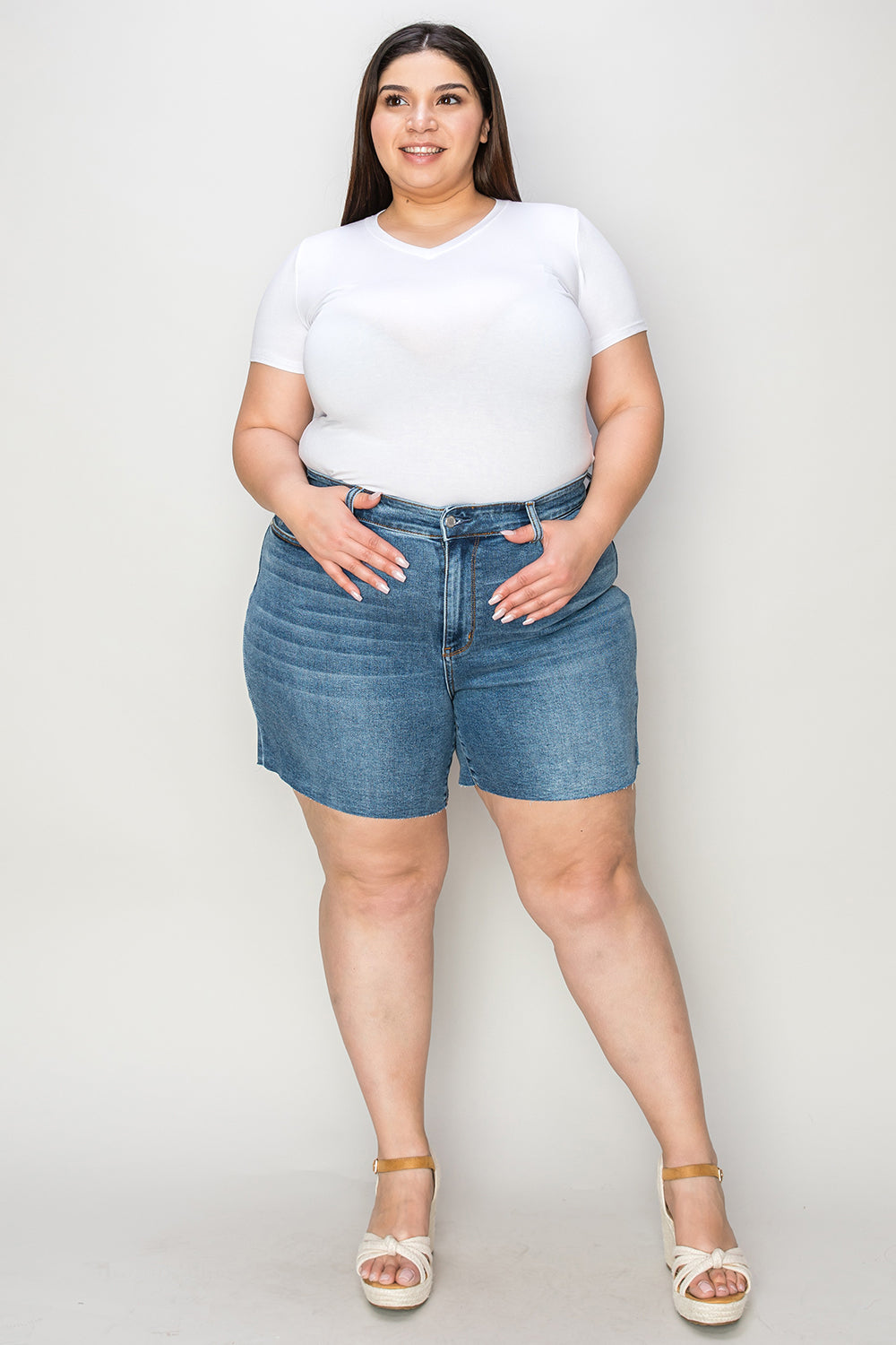 A woman in a white short-sleeve top and Judy Blue Full Size High Waist Slim Denim Shorts stands against a plain backdrop. She is smiling with her hands resting on her hips, wearing white open-toe wedge sandals that emphasize the quality denim fabric of her high-waisted design.