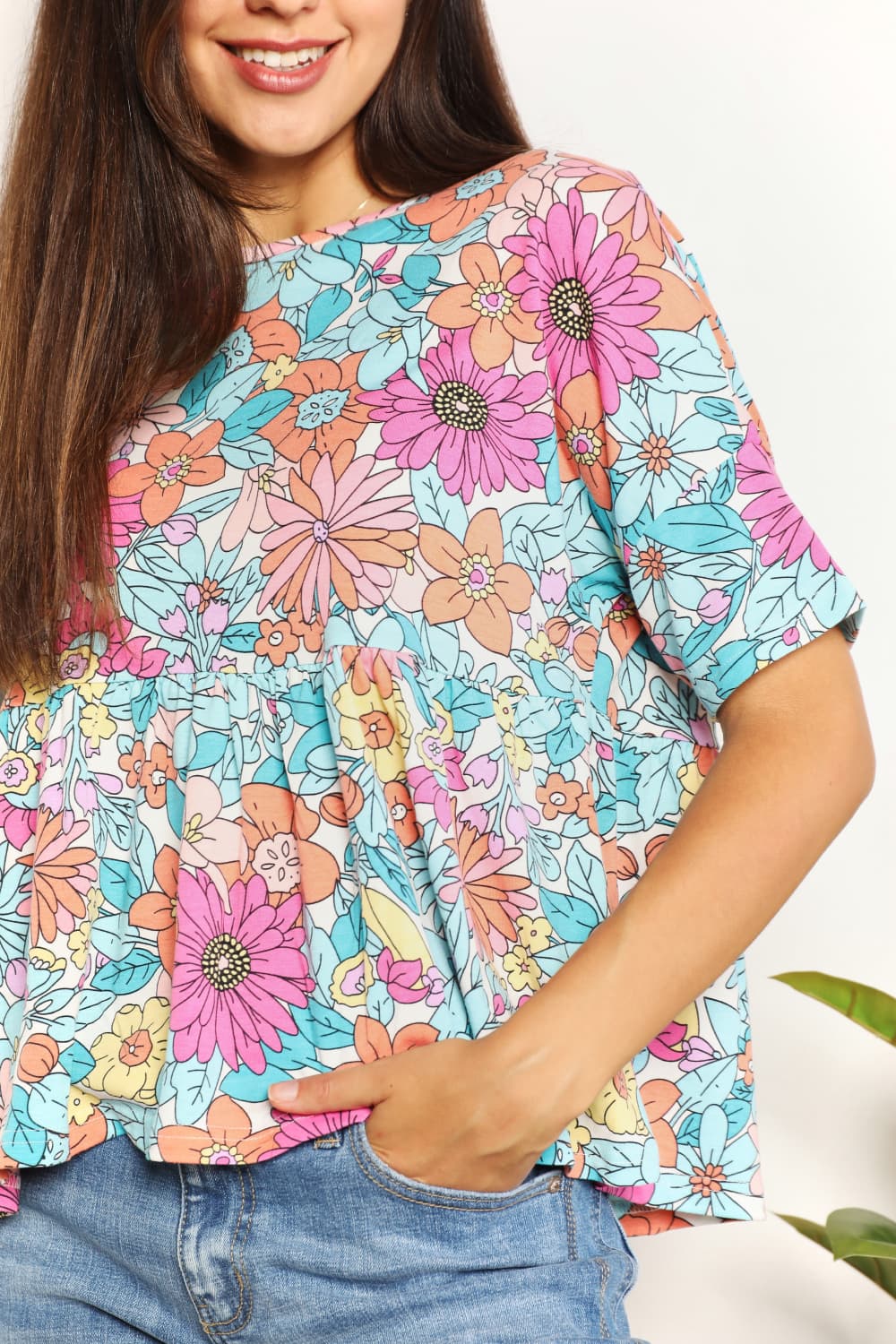 A person wearing a Double Take Floral Round Neck Babydoll Top and jeans stands with one hand in their pocket, with a plant in the background.