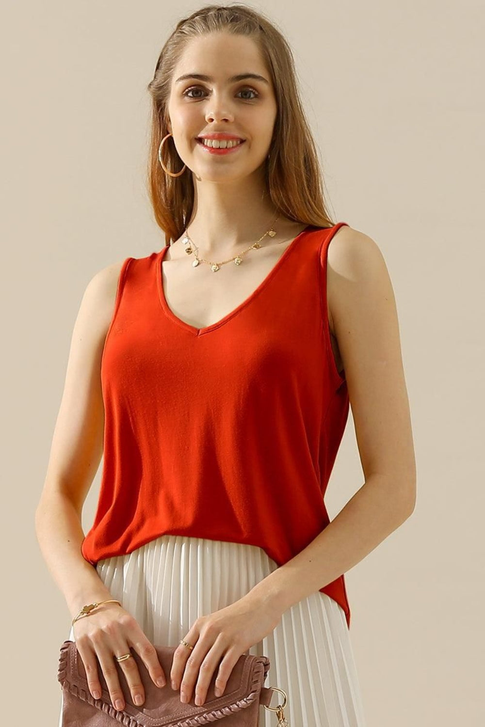 A woman in a stylish and versatile Ninexis Full Size V-Neck Curved Hem Tank and white pleated skirt poses while holding her handbag.