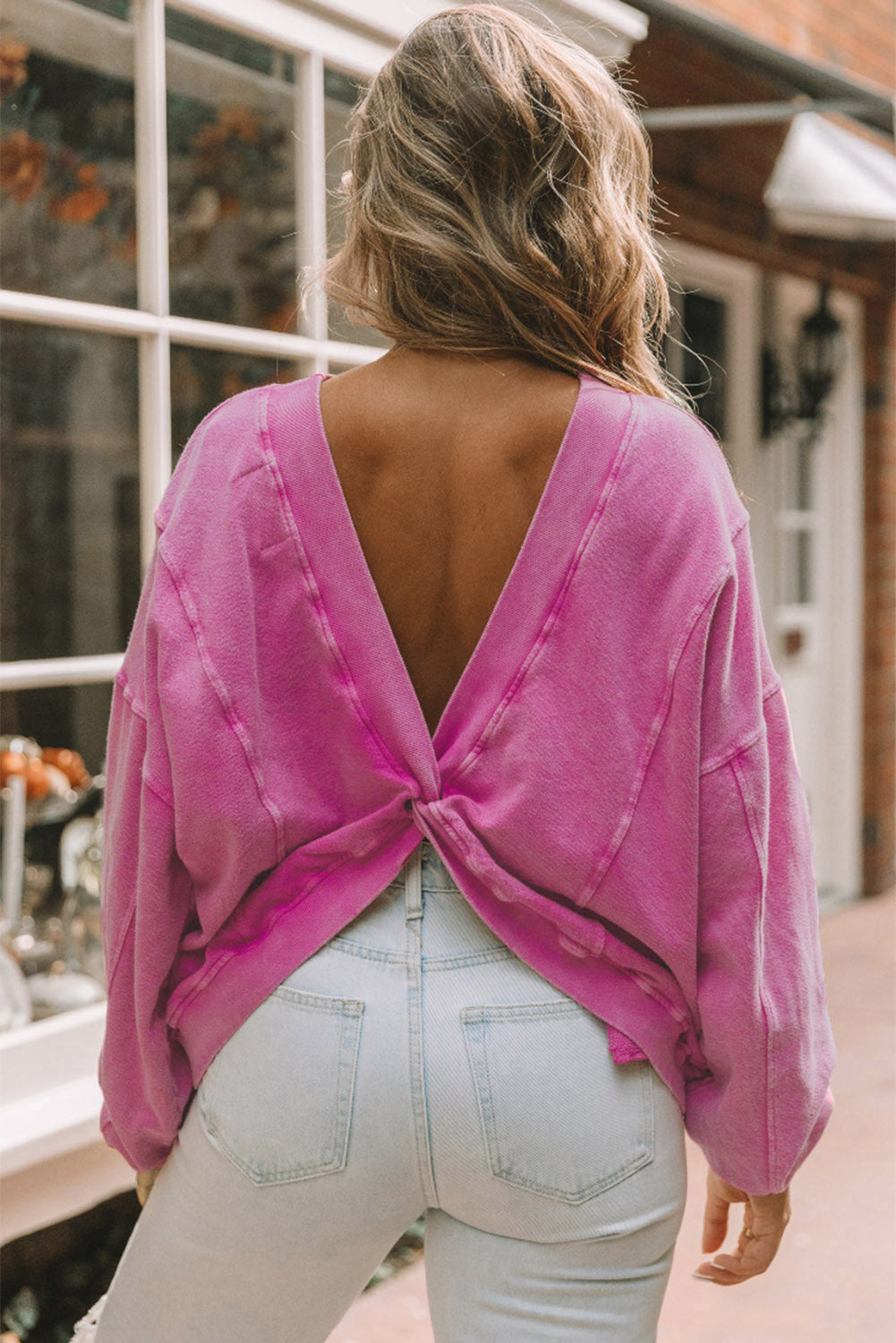 A woman stands with her back to the camera, wearing a long-sleeved Rose Exposed Seam Twist Open Back Oversized Sweatshirt and light blue jeans, showcasing a relaxed fit.