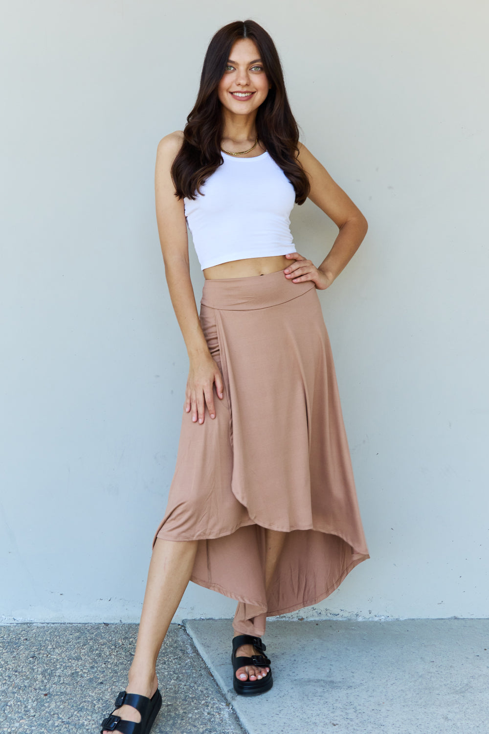 Person wearing a white crop top, the Ninexis First Choice High Waisted Flare Maxi Skirt in Camel with an asymmetrical hemline, and black sandals, standing against a light-colored wall.