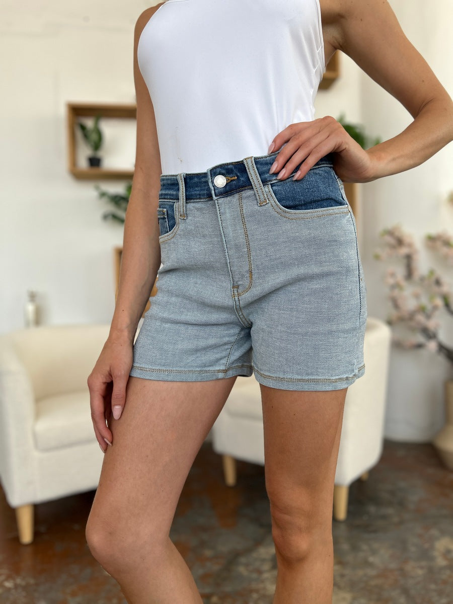 A person models indoors beside a beige chair and a plant, sporting a white t-shirt with the Judy Blue Full Size Color Block Denim Shorts for an effortlessly bold, eye-catching look.
