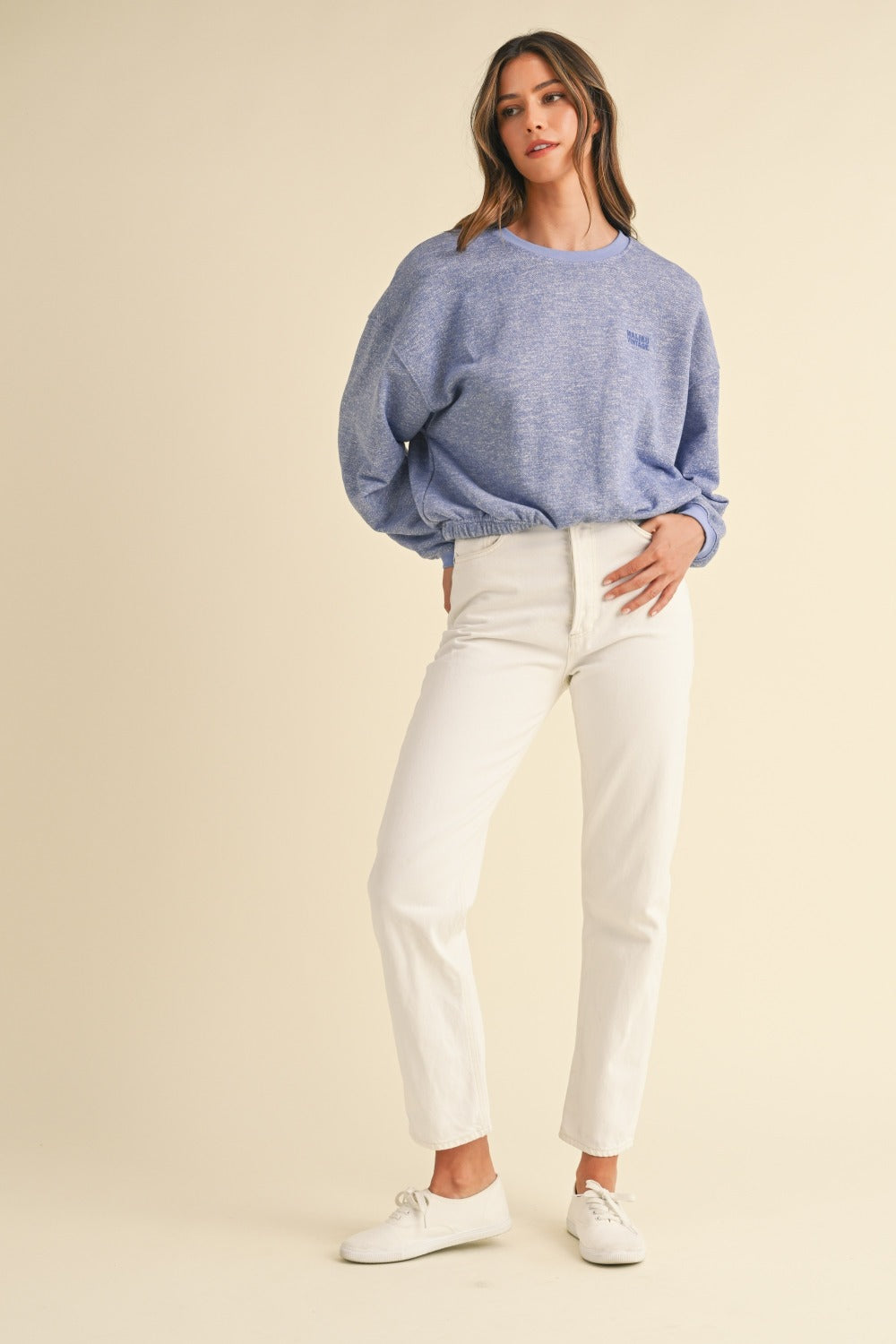 A person wearing a Mable Round Neck Letter Embroidery Crop Sweatshirt in blue, paired with white pants, stands against a plain background, touching the collar of the sweatshirt with one hand.