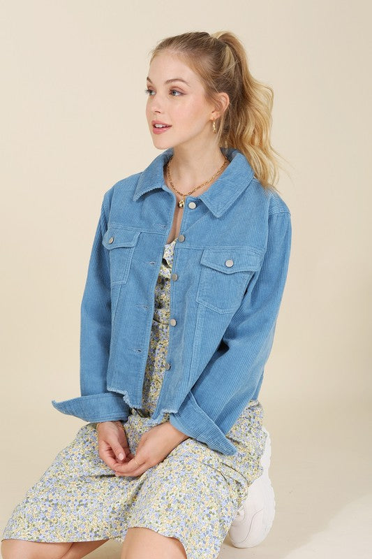 A person with long hair wears a blue frayed corduroy jacket over a floral dress, seated on a light-colored background.